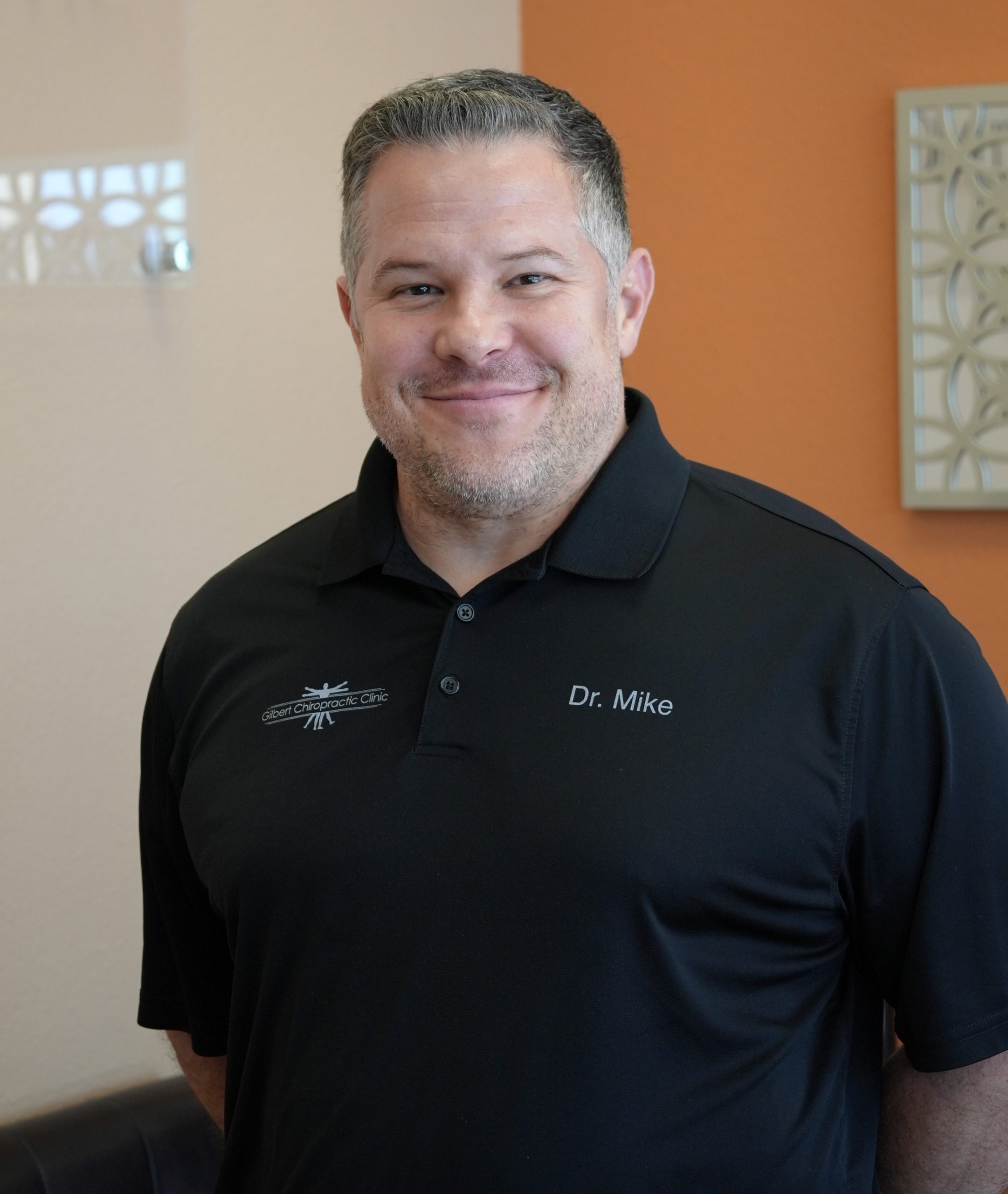 Michael Abromovitz standing in front of orange wall smiling with a black polo shirt