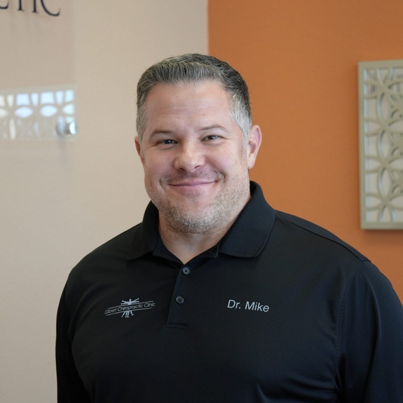 Chiropractor Gilbert AZ Michael Abromovitz Michael Abromovitz standing in front of orange wall smiling with a black polo shirt